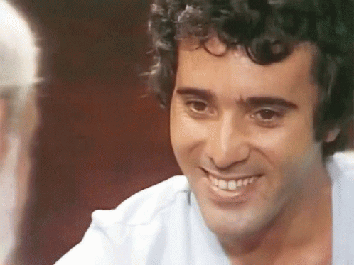 A Man With Curly Hair Is Smiling And Talking To Another Man . GIF