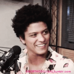 A Man With Curly Hair Is Smiling In Front Of A Microphone GIF