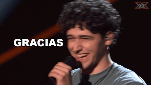 A Man With Curly Hair Is Smiling In Front Of A Sign That Says " Gracias " GIF