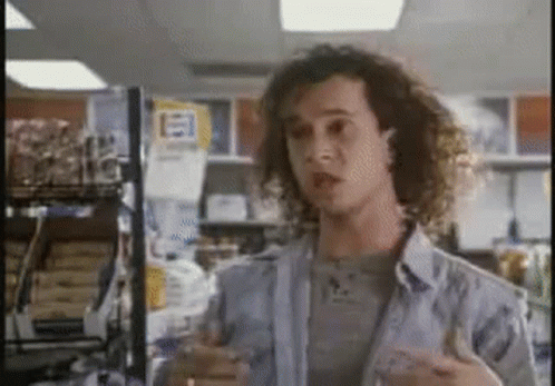 A Man With Curly Hair Is Standing In Front Of A Sign That Says ' E ' On It . GIF