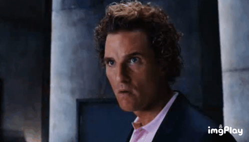 A Man With Curly Hair Is Wearing A Suit And A Pink Shirt And Making A Funny Face . GIF