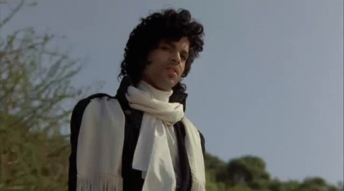 A Man With Curly Hair Is Wearing A White Scarf Around His Neck And A Black Jacket . GIF