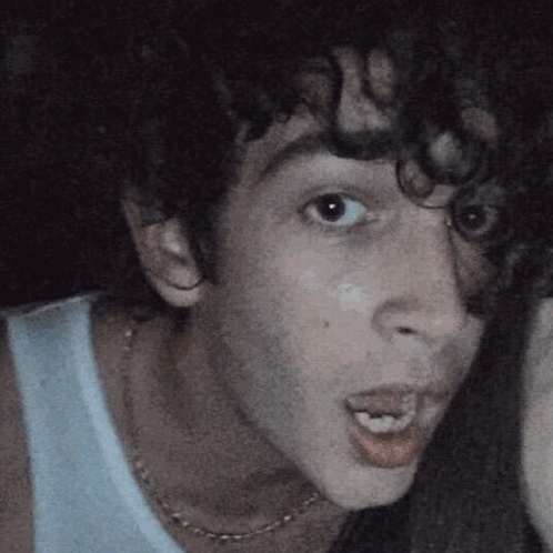 A Man With Curly Hair Is Wearing A White Tank Top And A Necklace GIF