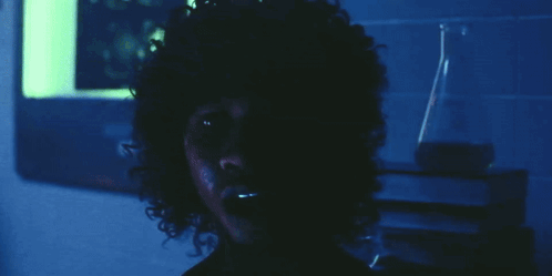 A Man With Curly Hair Looks At The Camera In A Dark Room GIF