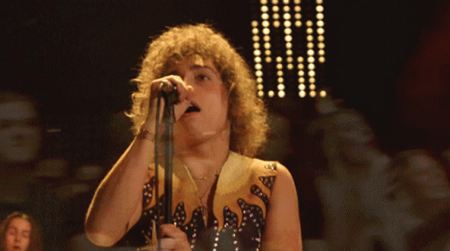 A Man With Curly Hair Singing Into A Microphone While Wearing A Black And Gold Outfit GIF