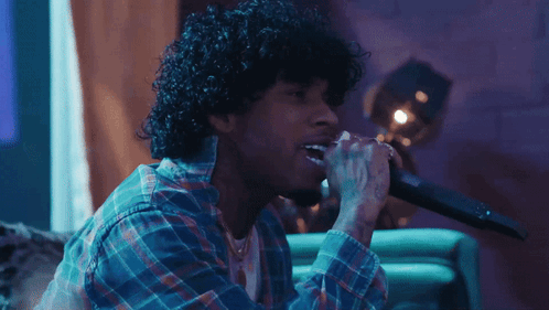 A Man With Curly Hair Singing Into A Microphone While Wearing A Plaid Shirt GIF