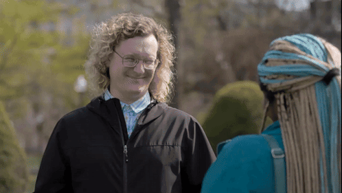 A Man With Curly Hair Wearing Glasses And A Black Jacket Talks To A Woman With Braids GIF