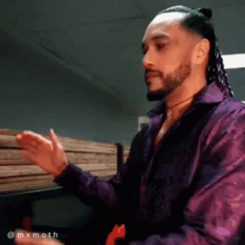 A Man With Dreadlocks And A Beard Wearing A Purple Shirt Is Standing In Front Of A Stack Of Wood GIF