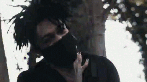 A Man With Dreadlocks And A Black Mask On His Face Is Standing In Front Of A Tree . GIF