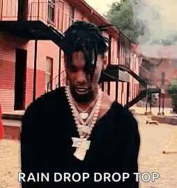 A Man With Dreadlocks And A Necklace Is Standing In Front Of A Building Smoking A Cigarette . GIF