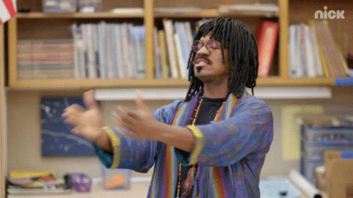 A Man With Dreadlocks And Glasses Is Standing In Front Of A Bookshelf With The Nick Logo On It GIF