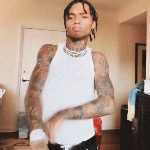 A Man With Dreadlocks And Tattoos Is Wearing A White Tank Top And Black Pants . GIF