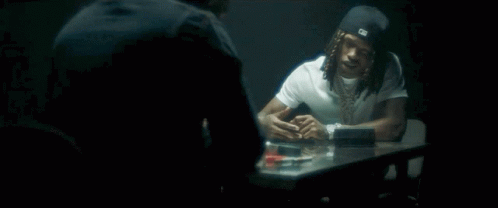 A Man With Dreadlocks Is Sitting At A Table Talking To Another Man In A Dark Room . GIF