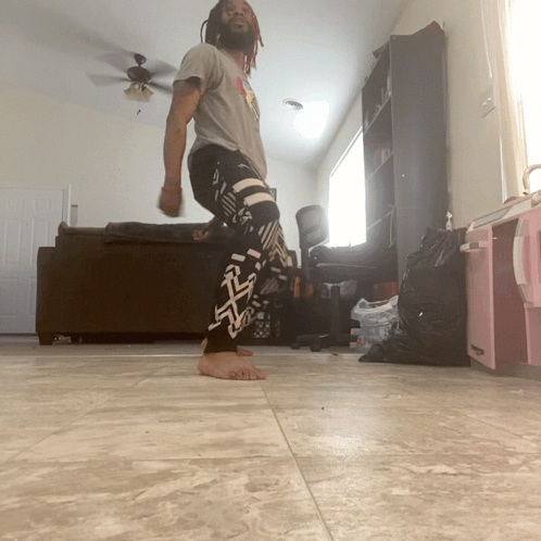 A Man With Dreadlocks Is Standing In A Living Room Wearing A Pair Of Black And White Pants With The Letter R On Them GIF