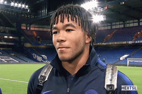 A Man With Dreadlocks Is Standing In Front Of A Stadium That Says Heitse GIF