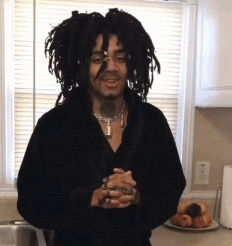 A Man With Dreadlocks Is Standing In Front Of A Window Holding A Glass GIF