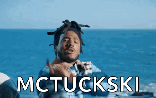 a man with dreadlocks is standing in front of the ocean and the words mctuckski are displayed above him