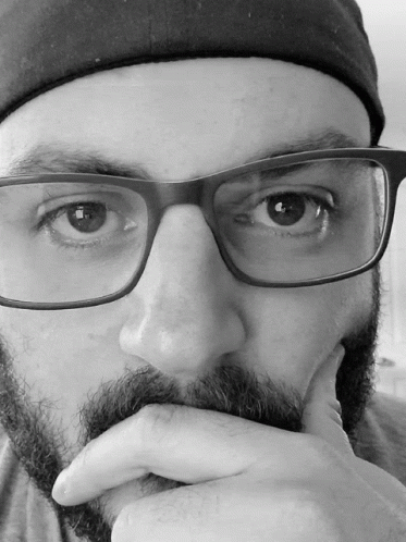 A Man With Glasses And A Beard Holds His Hand To His Face GIF