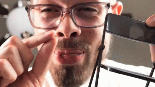 A Man With Glasses And A Beard Is Holding A Black Object In Front Of His Face GIF