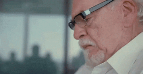 A Man With Glasses And A Beard Is Looking Down At Something . GIF
