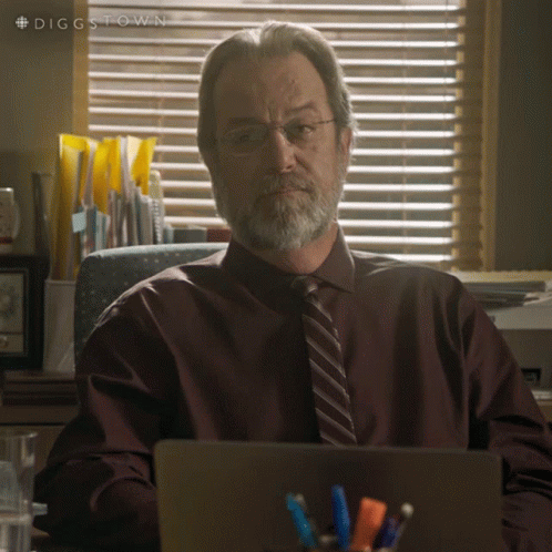 A Man With Glasses And A Beard Is Sitting At A Desk With A Laptop In Front Of Him GIF