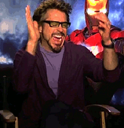 A Man With Glasses And A Beard Is Sitting In Front Of A Man In A Superhero Costume GIF