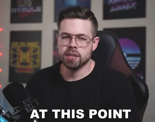 A Man With Glasses And A Beard Is Sitting In Front Of A Microphone With The Words At This Point . GIF