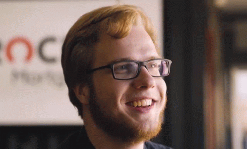 A Man With Glasses And A Beard Is Smiling In Front Of A Sign That Says Oc GIF