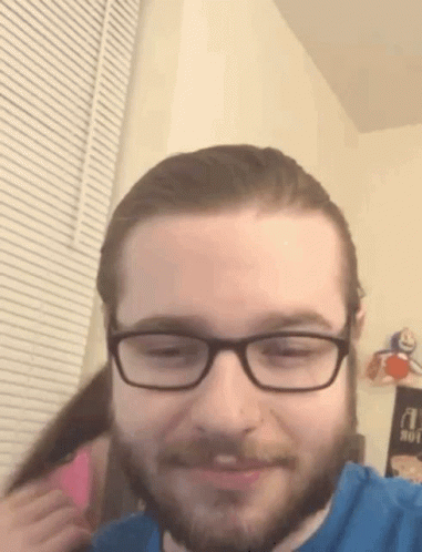 A Man With Glasses And A Beard Is Smiling While A Woman Holds His Hair . GIF