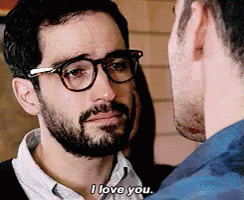A Man With Glasses And A Beard Is Talking To Another Man Who Is Wearing A Blue Shirt . GIF