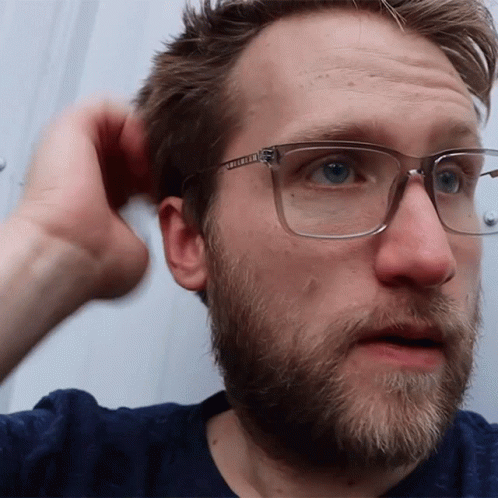 A Man With Glasses And A Beard Looks At The Camera With His Hand In His Hair GIF