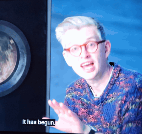 A Man With Glasses And A Blue Sweater Says It Has Begun On A Screen GIF