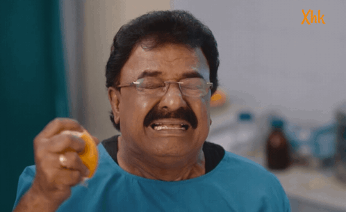 A Man With Glasses And A Mustache Is Holding An Orange In Front Of A Sign That Says Xmk GIF