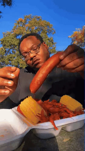 A Man With Glasses Is Eating Corn On The Cob And Crayfish GIF