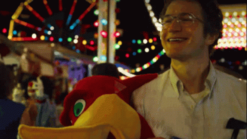 A Man With Glasses Is Holding A Stuffed Bird At A Carnival GIF