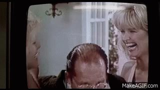 A Man With Glasses Is Laughing While A Woman Laughs Behind Him On A Television Screen . GIF
