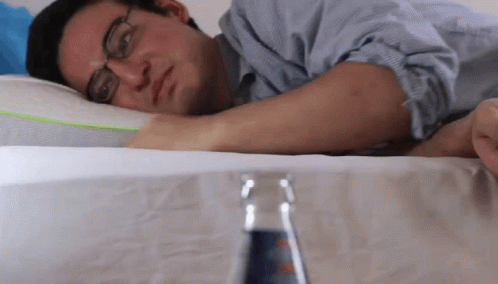 A Man With Glasses Is Laying On A Bed With A Bottle Of Soda In Front Of Him GIF