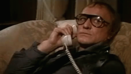 A Man With Glasses Is Sitting On A Couch Talking On A Phone . GIF