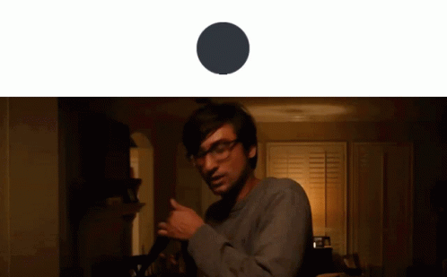 A Man With Glasses Is Standing In A Dark Room With A Black Circle Behind Him GIF