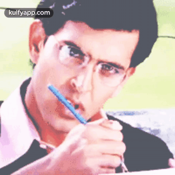 A Man With Glasses Is Writing On A Piece Of Paper With A Pencil In His Mouth . GIF
