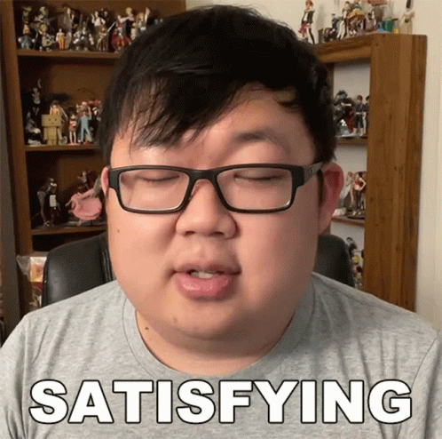 A Man With Glasses Says Satisfying In Front Of A Shelf Full Of Figurines GIF