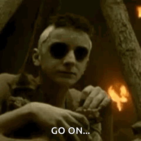A Man With Glowing Eyes Is Sitting In A Dark Room With A Fire In The Background And Says `` Go On '' . GIF