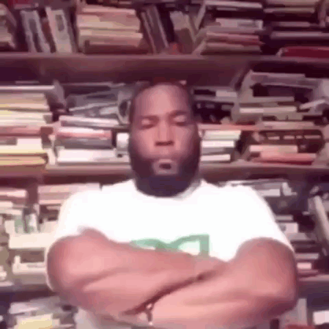 A Man With His Arms Crossed Is Sitting In Front Of A Bookshelf Filled With Books . GIF