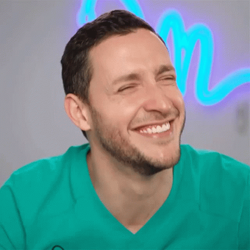 A Man With His Eyes Closed Is Smiling In Front Of A Neon Sign That Says M GIF
