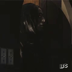 A Man With Long Black Hair And Glasses Is Standing In A Dark Room With A Sign That Says Ifc GIF