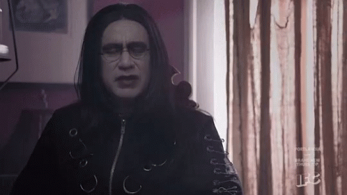 A Man With Long Black Hair And Glasses Is Standing In Front Of A Window In A Room . GIF