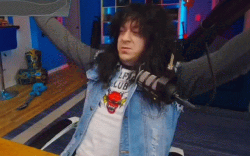 A Man With Long Black Hair Is Sitting At A Table With His Arms Outstretched And Holding A Microphone . GIF