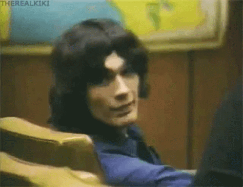 A Man With Long Black Hair Is Sitting In A Chair And Looking At The Camera . GIF