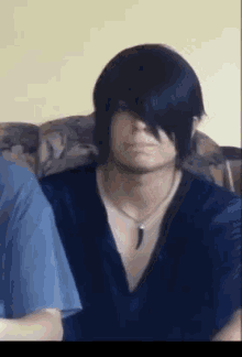A Man With Long Black Hair Is Sitting On A Couch Wearing A Blue Shirt And A Necklace . GIF