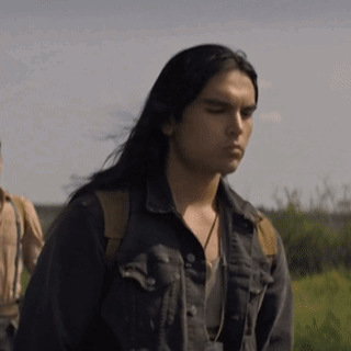 A Man With Long Black Hair Is Wearing A Denim Jacket And Standing In A Field . GIF